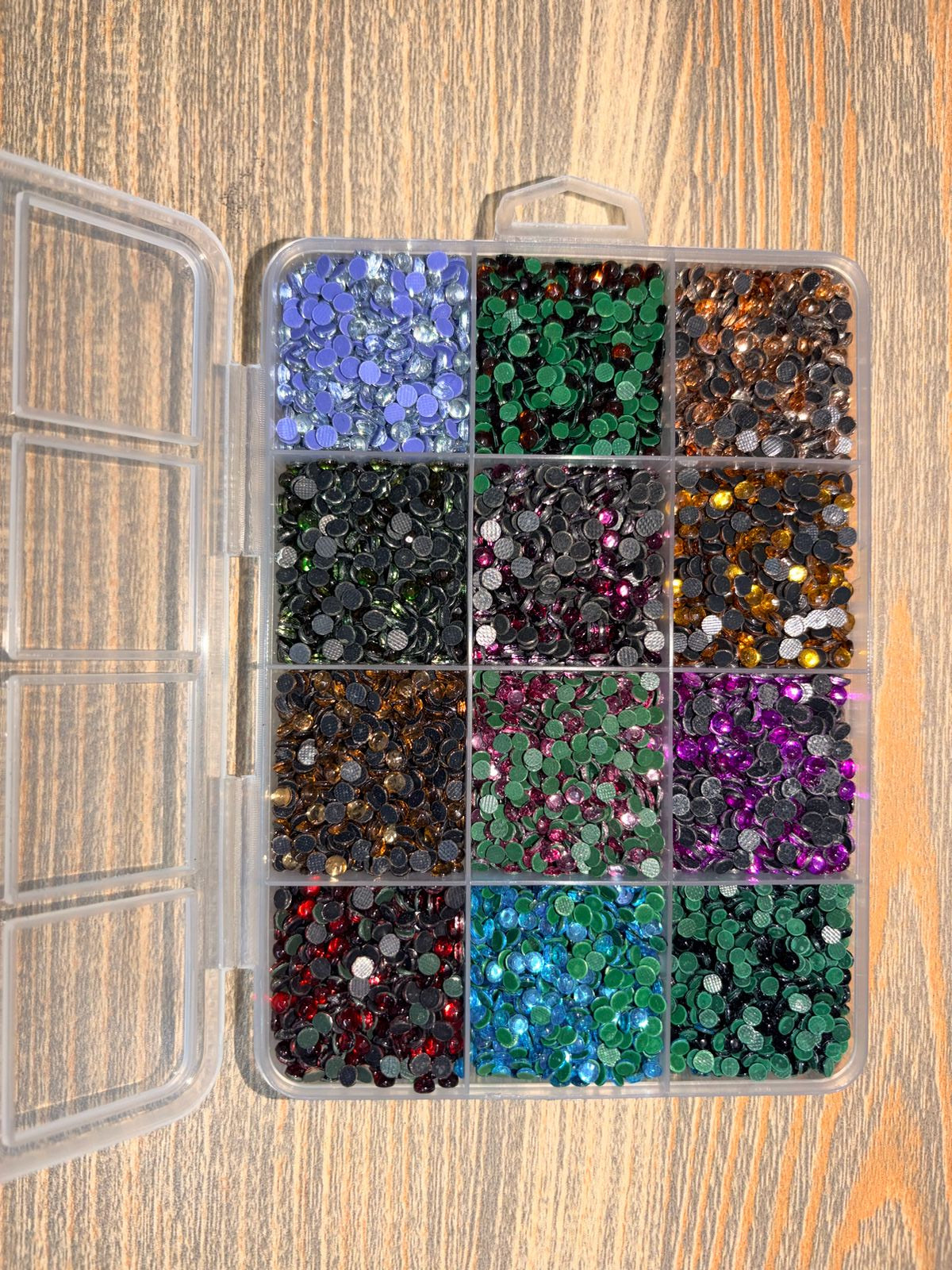 A clear plastic organizer box filled with many small round sequins or rhinestones in assorted colors, photographed from above on a wood surface.